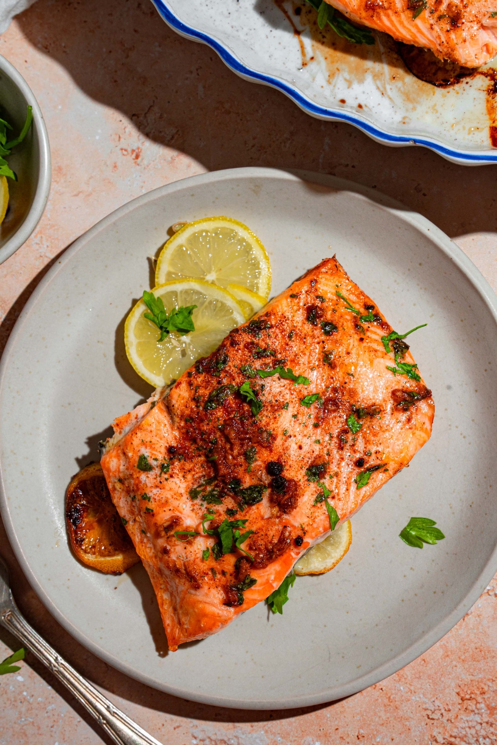 A white plate with baked Sockeye salmon garnished with fresh parsley and served with sliced lemon. The plate is on a tan counter with a baking dish with salmon and small bowl of garnishes.