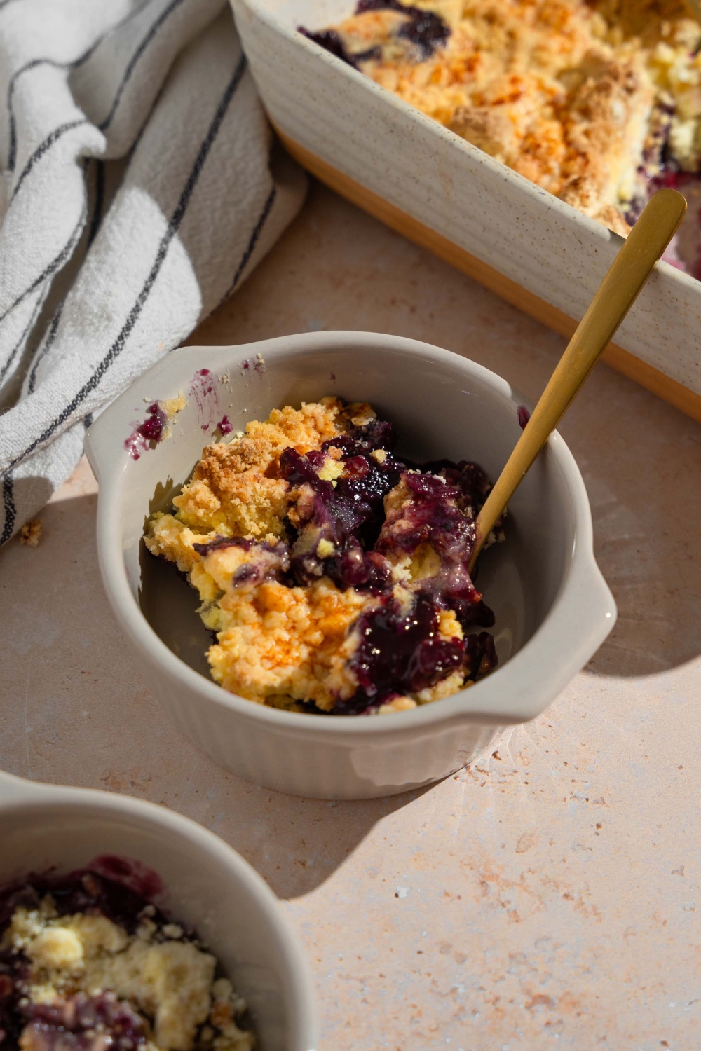 A ramekin with a bite of blueberry dump cake with a spoon in the cake. The ramekin is on a tan counter with a baking dish of dump cake and a white striped napkin.