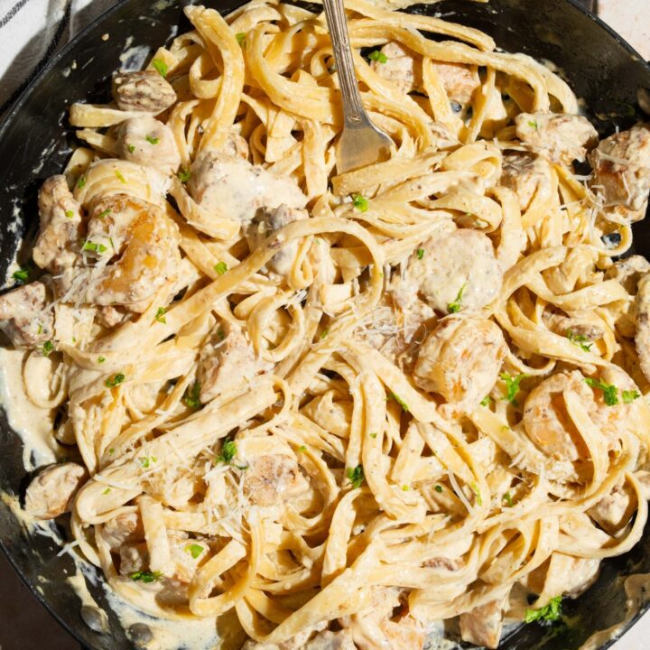 A skillet with a fork tossing chicken and shrimp alfredo garnished with fresh parsley. The skillet is on a tan counter with a white striped napkin.
