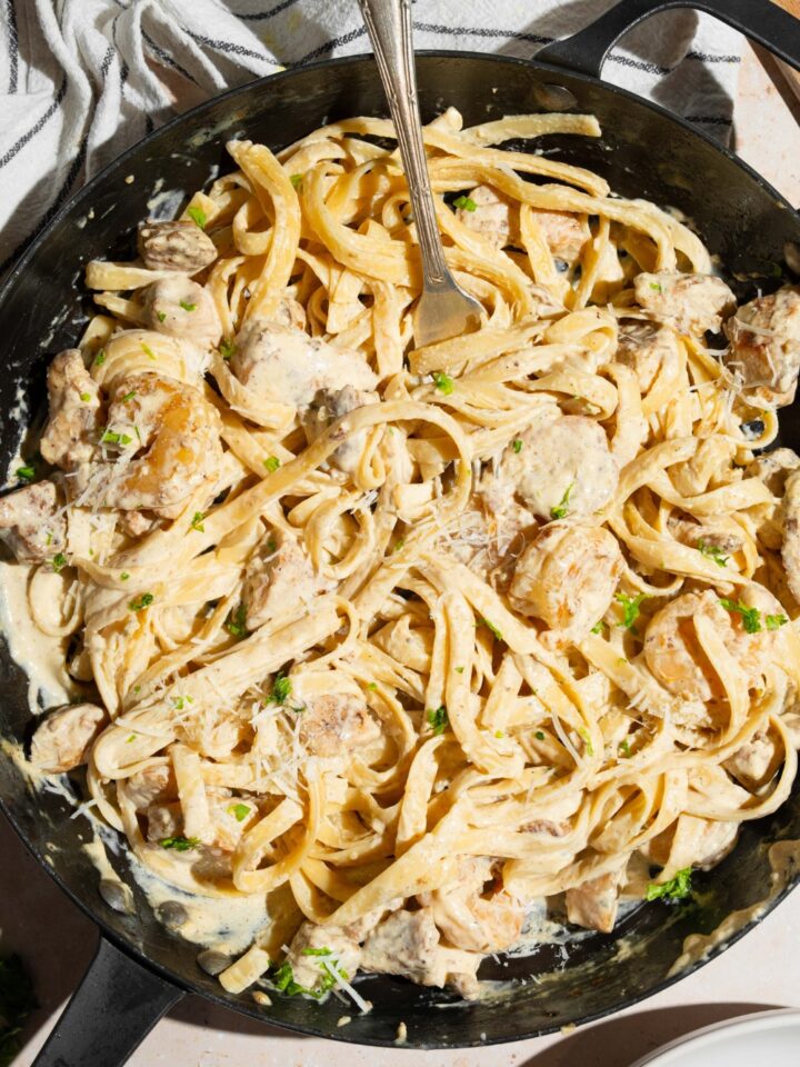 A skillet with a fork tossing chicken and shrimp alfredo garnished with fresh parsley. The skillet is on a tan counter with a white striped napkin.