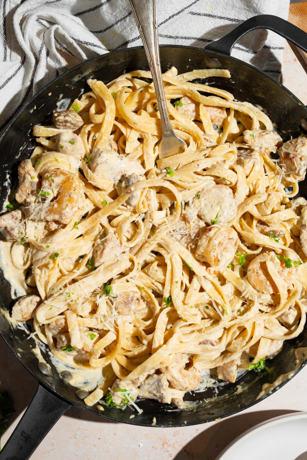 A skillet with a fork tossing chicken and shrimp alfredo garnished with fresh parsley. The skillet is on a tan counter with a white striped napkin.