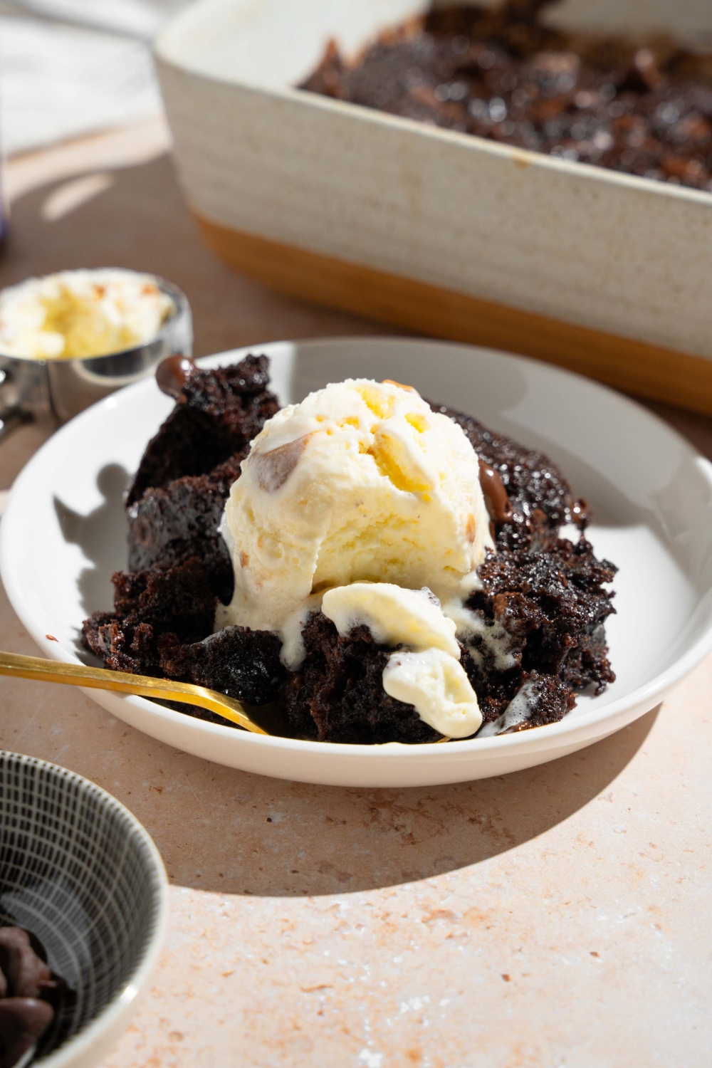 A white plate with a slice of chocolate dump cake topped with vanilla ice cream. There is a spoon on the plate. The plate is on a tan counter with a baking dish with dump cake.