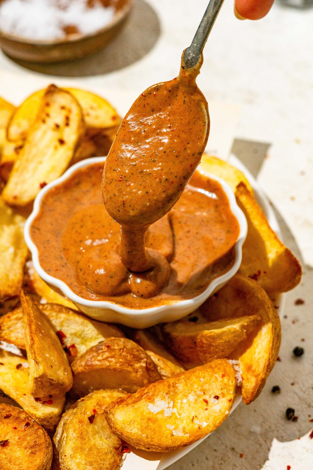 A small bowl of copycat Zaxby's sauce on a plate with wedge fries. There is a spoon dipping into the sauce. The plate is on a tan counter with a small bowl of salt.
