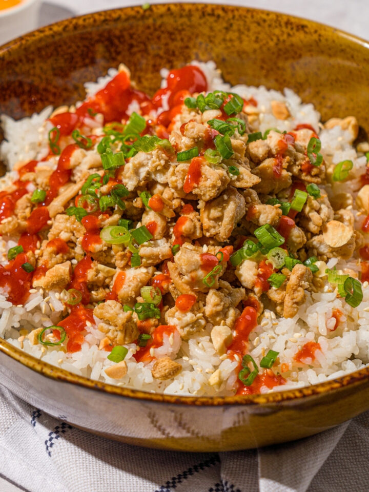 A bowl with ground turkey rice bowls with white rice, seasoned ground turkey, chopped peanuts, sliced green onions, sesame seeds, and drizzled with sriracha sauce. The bowl is on a white counter with a white striped napkin.