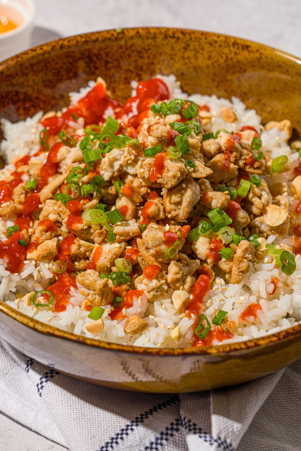 A bowl with ground turkey rice bowls with white rice, seasoned ground turkey, chopped peanuts, sliced green onions, sesame seeds, and drizzled with sriracha sauce. The bowl is on a white counter with a white striped napkin.