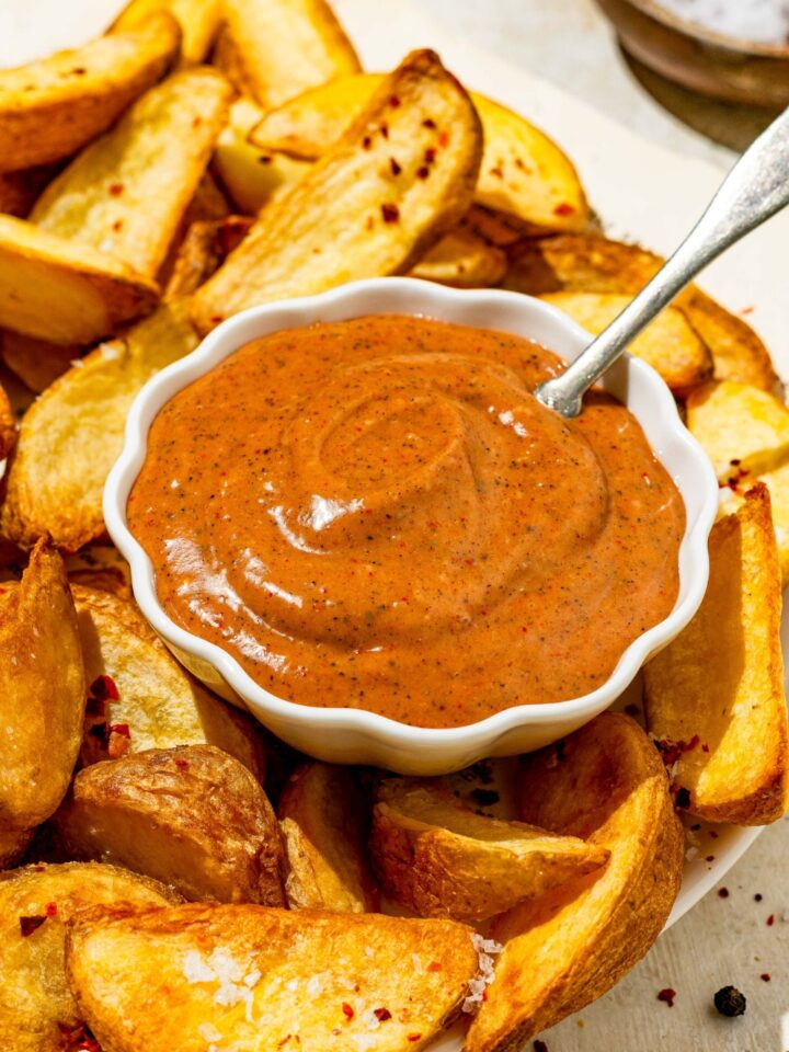 A small bowl of copycat Zaxby's sauce on a plate with wedge fries. There is a spoon in the sauce. The plate is on a tan counter with a small bowl of salt.