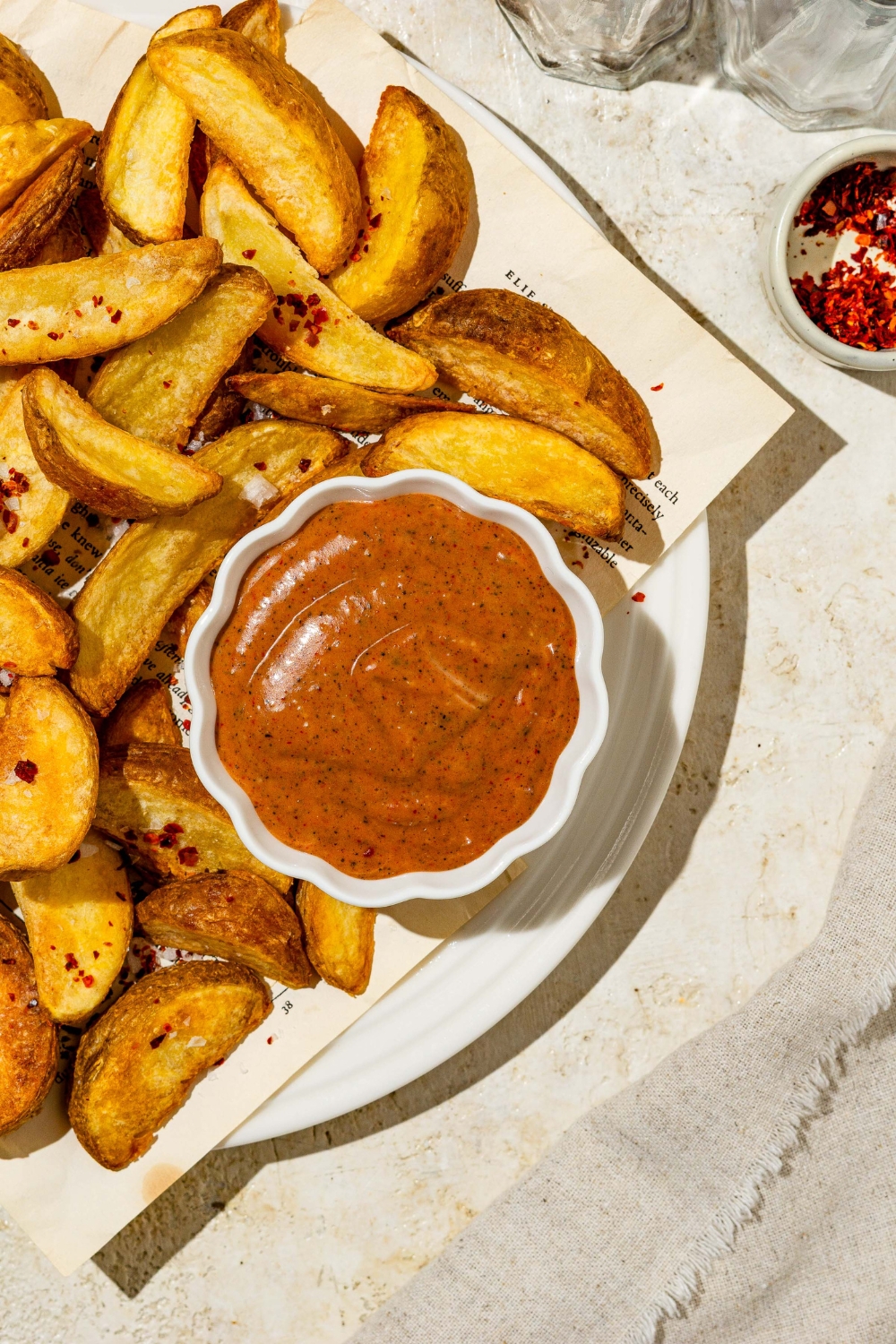 A small of Zaxby's sauce on a paper lined plate with wedge fries. The plate is on a tan counter with a small bowl of garnishes.