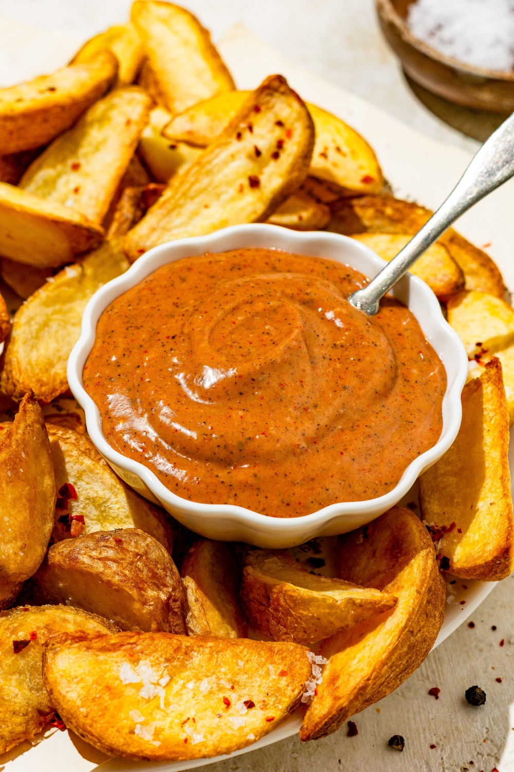 A small bowl of copycat Zaxby's sauce on a plate with wedge fries. There is a spoon in the sauce. The plate is on a tan counter with a small bowl of salt.
