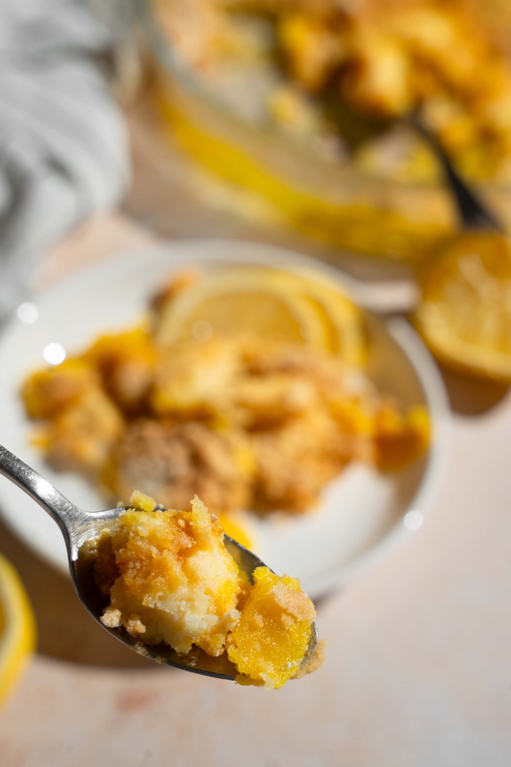 A close up of a spoon with a bite of lemon cream cheese dump cake. There is a plate with a slice of cake on a tan counter blurred in the background.