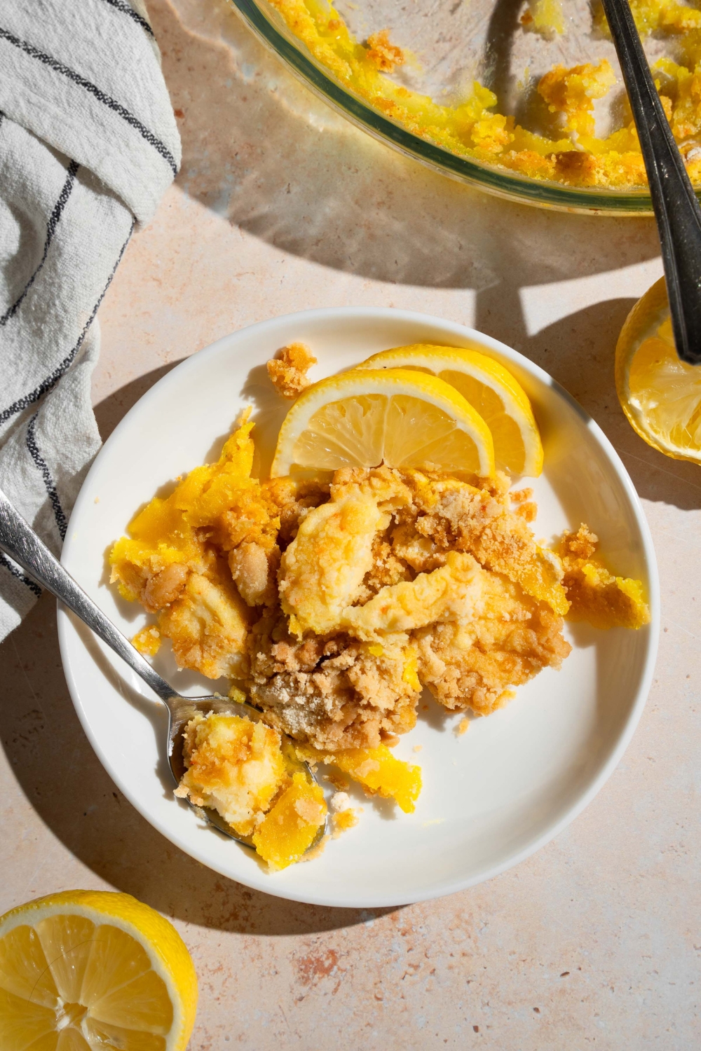 A white plate with a slice of lemon cream cheese dump cake served with sliced lemon and a fork. The plate is on a tan counter with a baking dish with cake and a white striped napkin.
