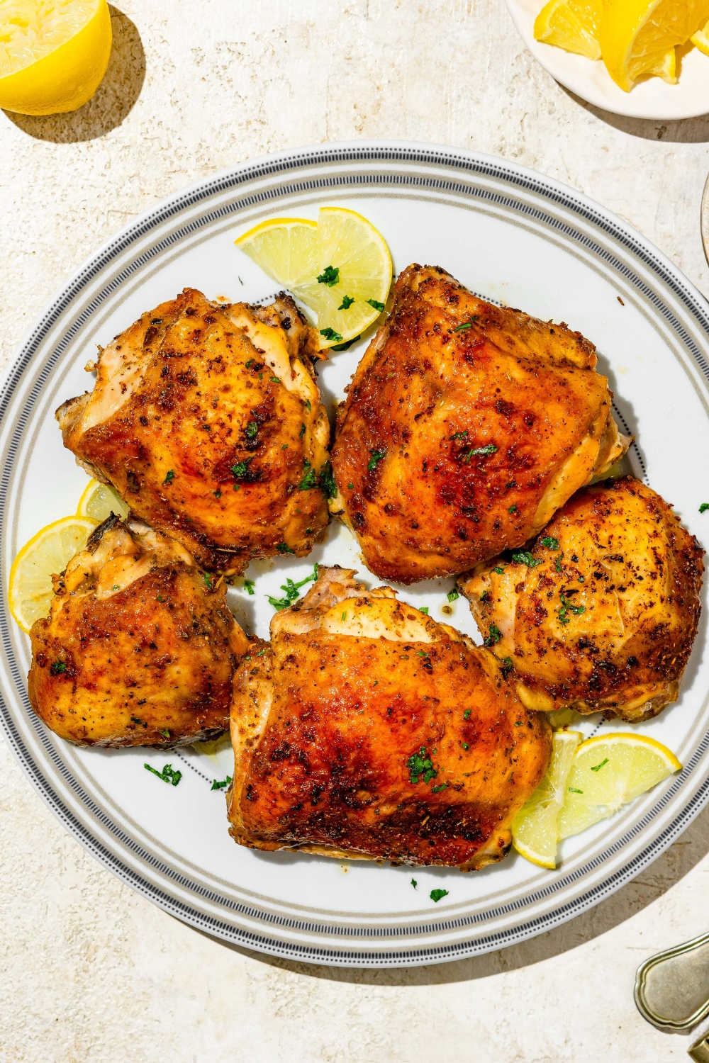 A plate of lemon pepper chicken thighs garnished with fresh parsley and lemon. The plate is on a tan counter with lemon.