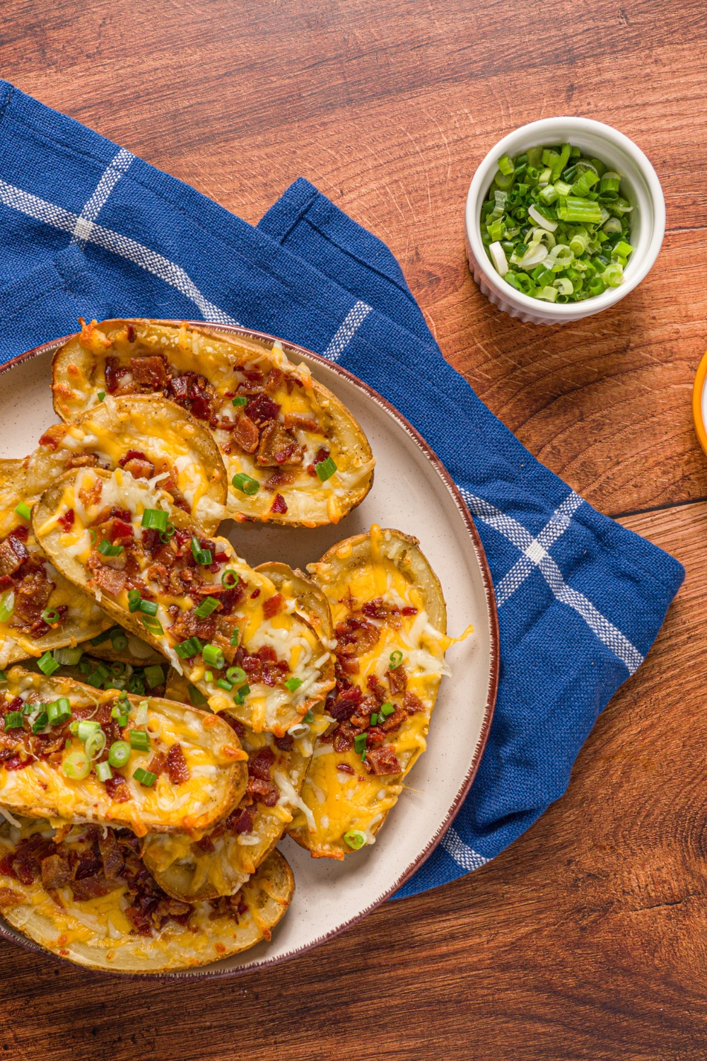 A plate with loaded potato skins garnished with sliced green onions. The plate is on a wooden counter with a blue striped napkin and small bowl of garnishes.
