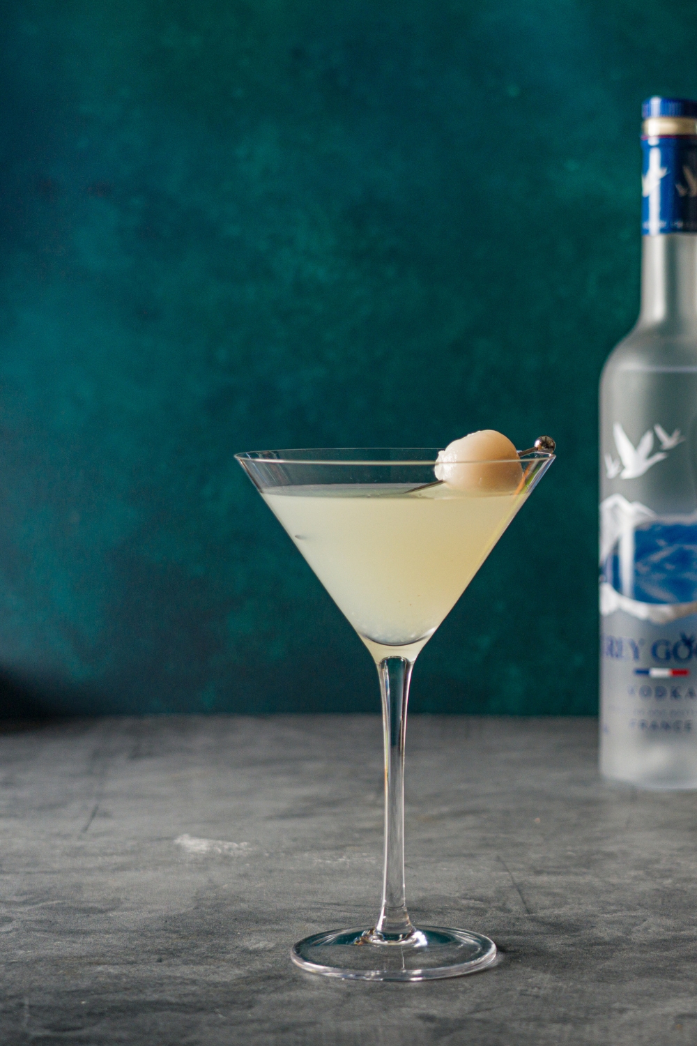 A lychee martini garnished with a lychee in a toothpick. The martini is on a stone counter with a bottle of Grey Goose.