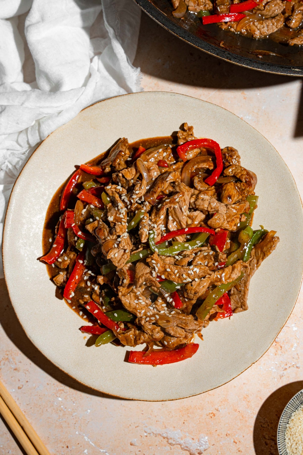 A plate of pepper steak tossed with stir fry and garnished with sesame seeds. The plate is on a tan counter with a white cloth napkin and pair of chopsticks.