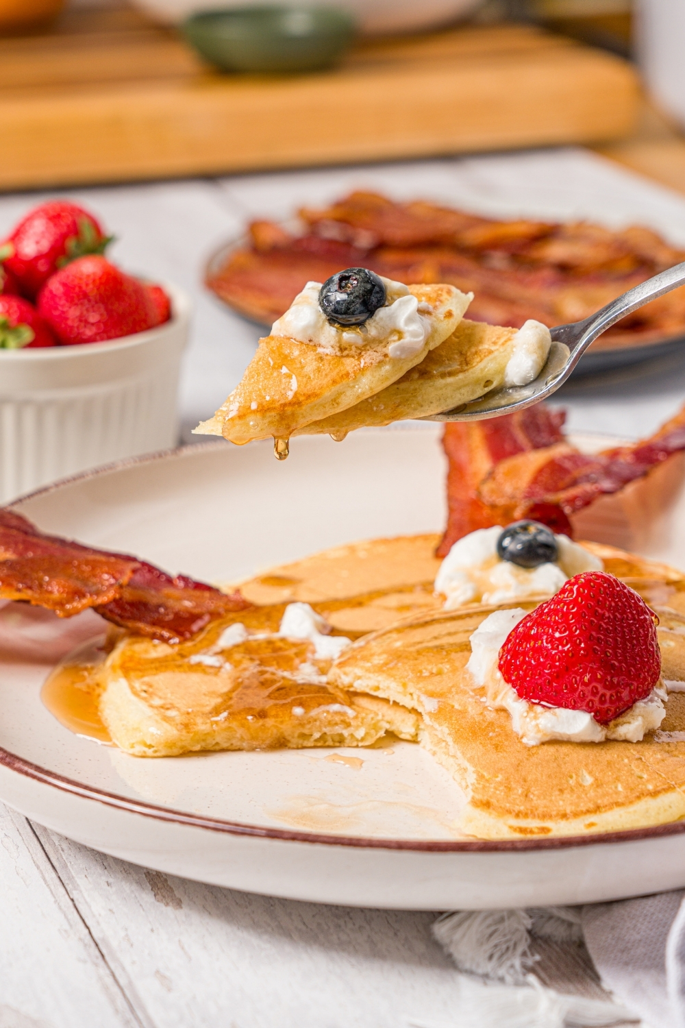 A fork taking a bite of reindeer pancakes from the plate. The plate is on a wood counter with a plate of bacon and bowl of berries.