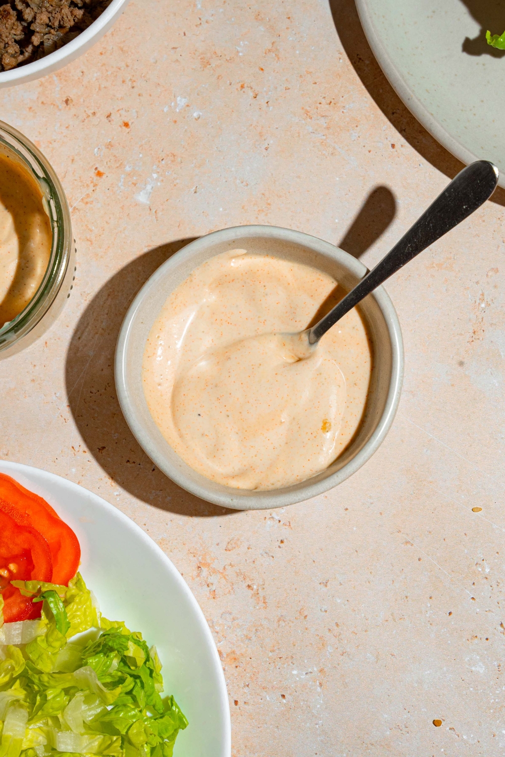 A bowl of smash burger sauce with a spoon in the bowl. The bowl is on a tan counter with a plate of burger toppings.