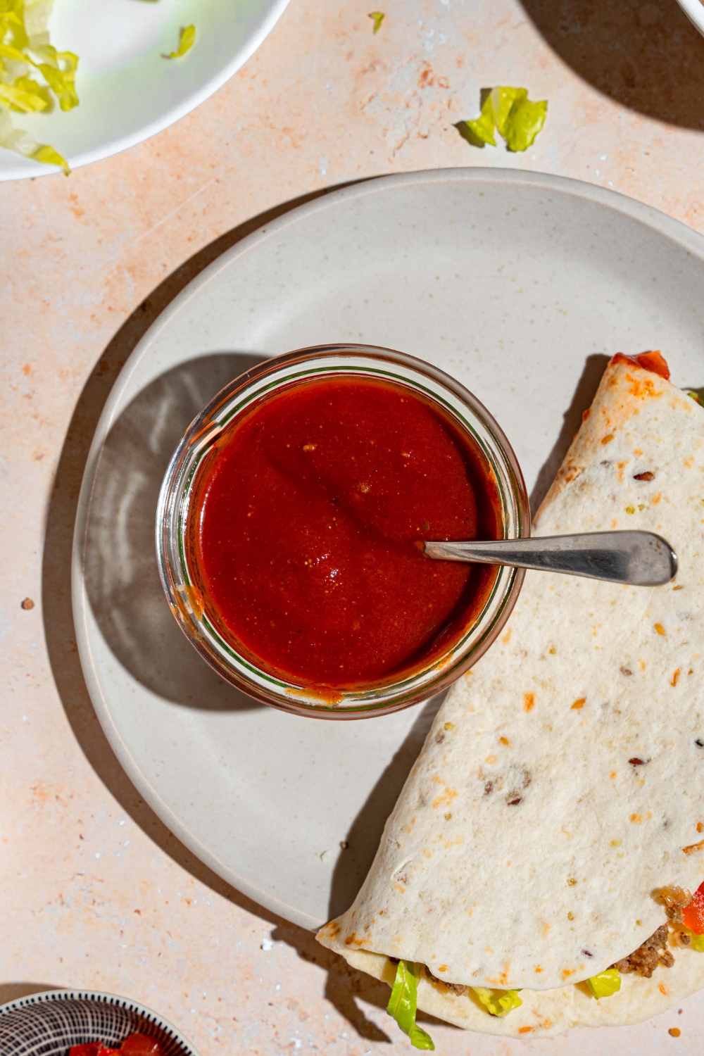 A glass jar of taco sauce with a spoon in the jar. The jar is on a white plate with a soft shell taco. The plate is on a tan counter with a plate of shredded lettuce.