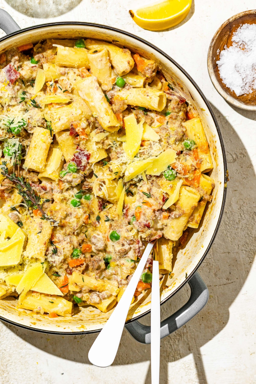 A dutch oven with rigatoni pasta with white bolognese. There is a fork and spoon in the pot. The dish is garnished with grated cheese. The dish is on a tan counter with a small bowl of salt and sliced lemon.