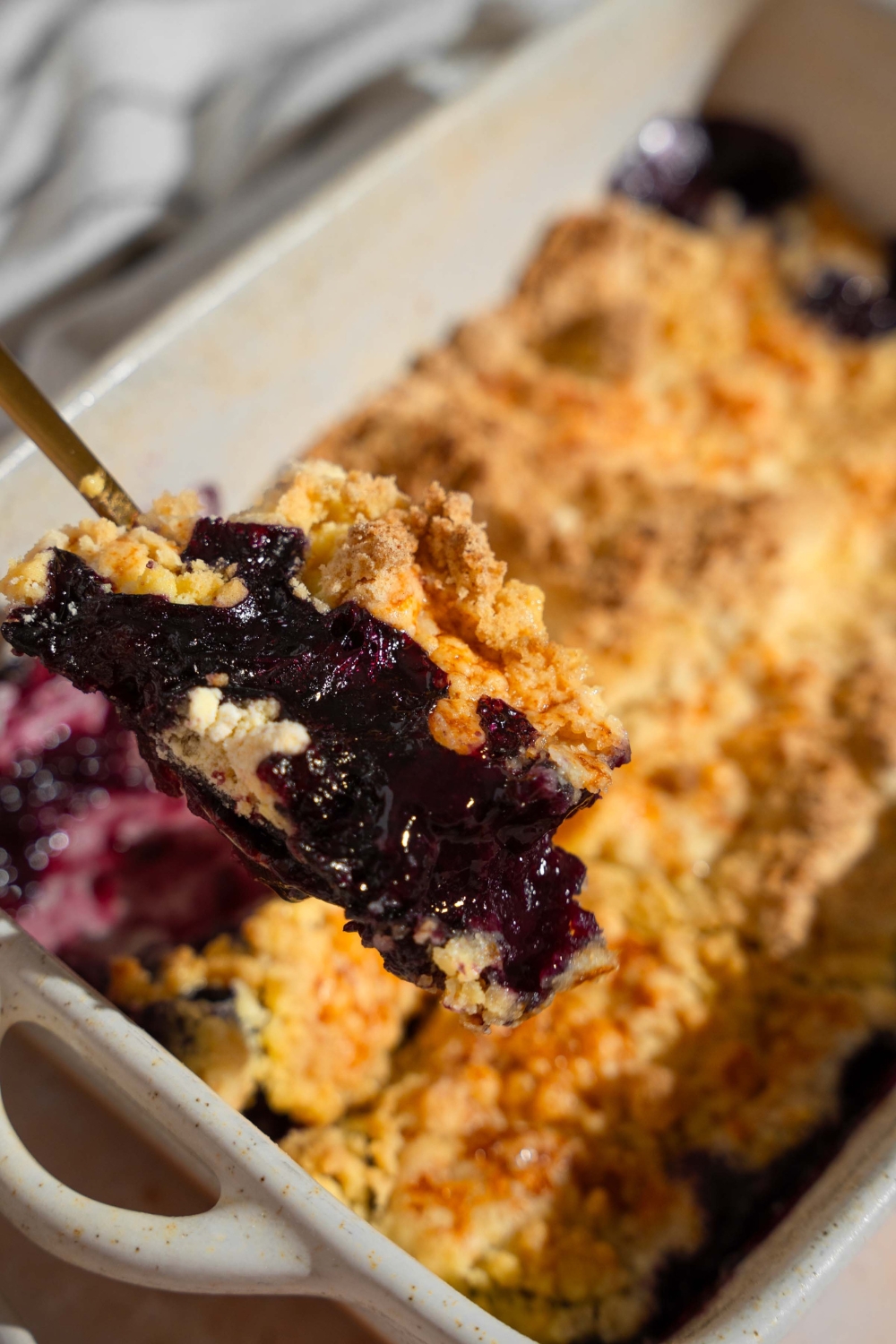 A close up of a fork with a bite of blueberry dump cake. There is a dish of cake blurred in the background.