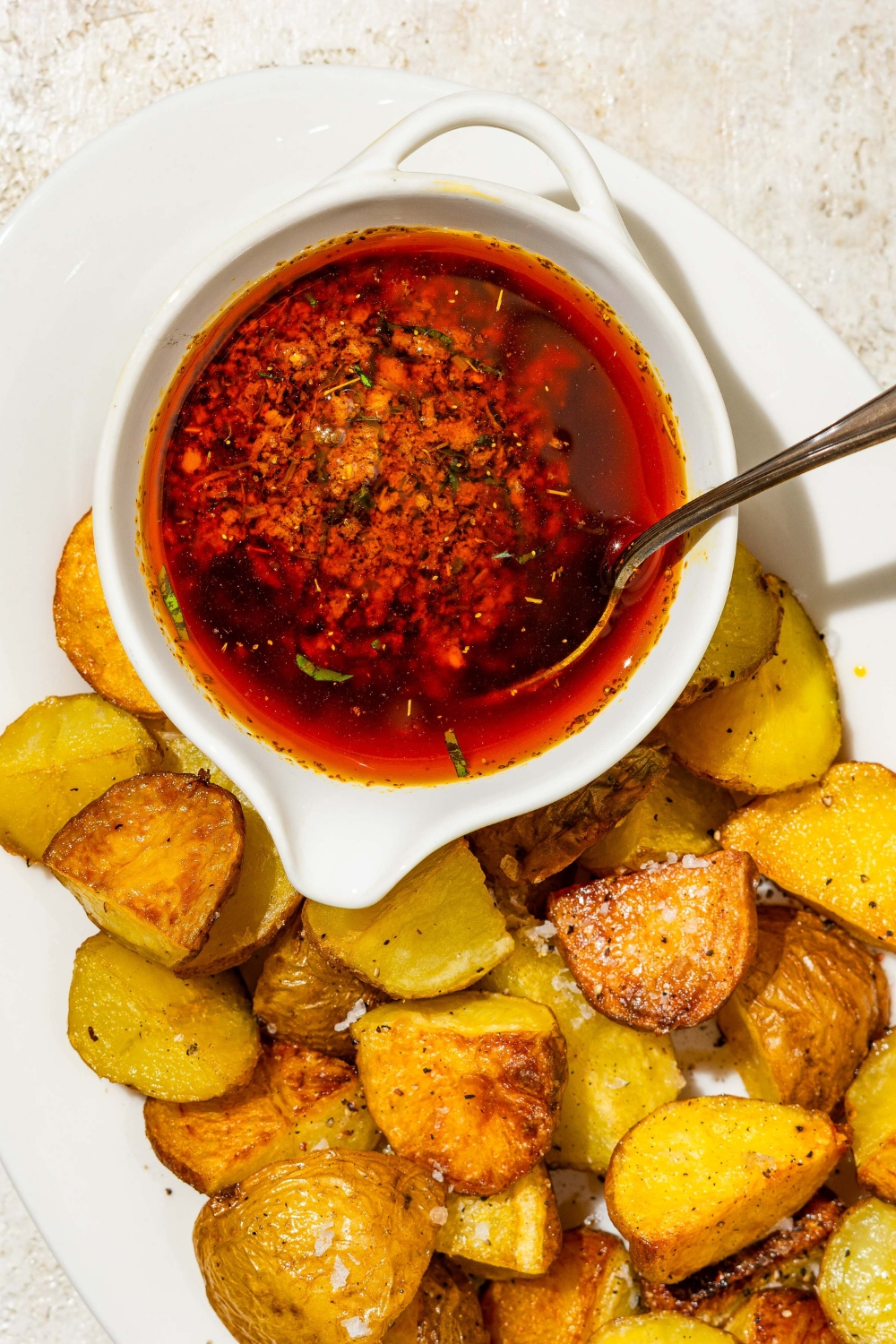A small bowl of cajun garlic butter sauce garnished with fresh parsley served on a plate with crispy potatoes. There is a spoon in the bowl. The plate is on a tan counter.