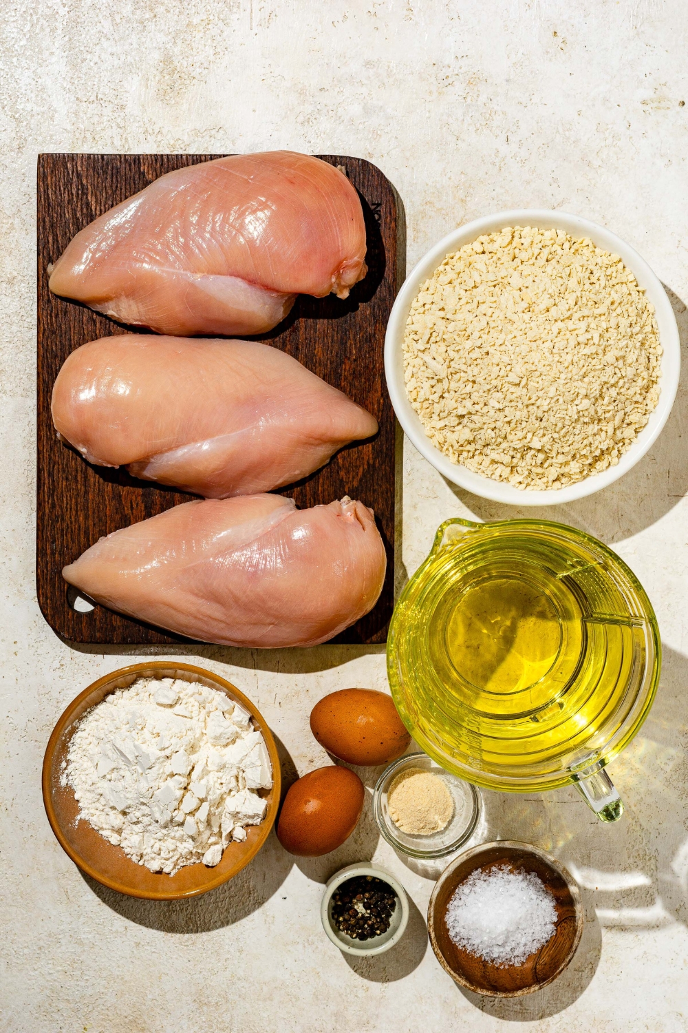 An overhead shot of several bowls in various sizes containing ingredients to make chicken Katsu including chicken breasts, panko breadcrumbs, eggs, flour, oil, and seasonings.