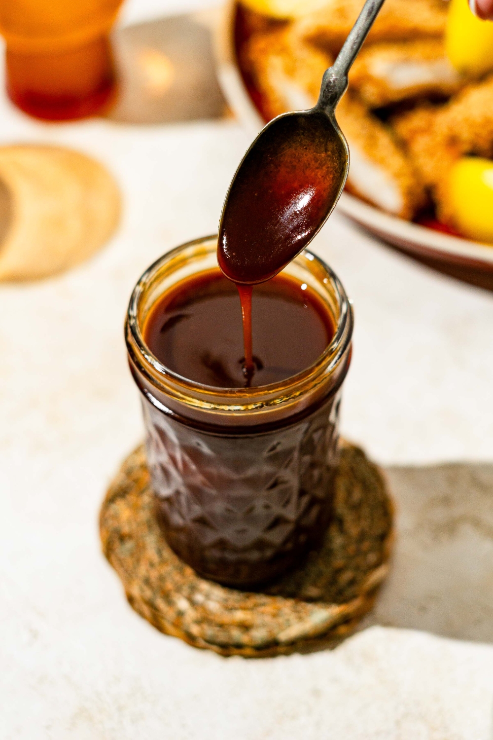 A mason jar with chicken Katsu sauce with a spoon dipped into the sauce. The jar is on a tan counter on a woven coaster.