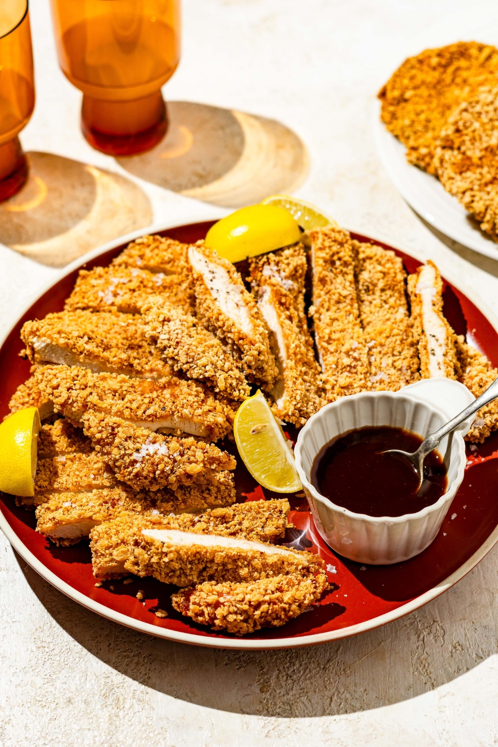 A plate of crispy chicken Katsu sliced in strips with a small ramekin of sauce with a spoon in the bowl. The plate is garnished with lemon wedges. The plate is on a tan counter with glasses.