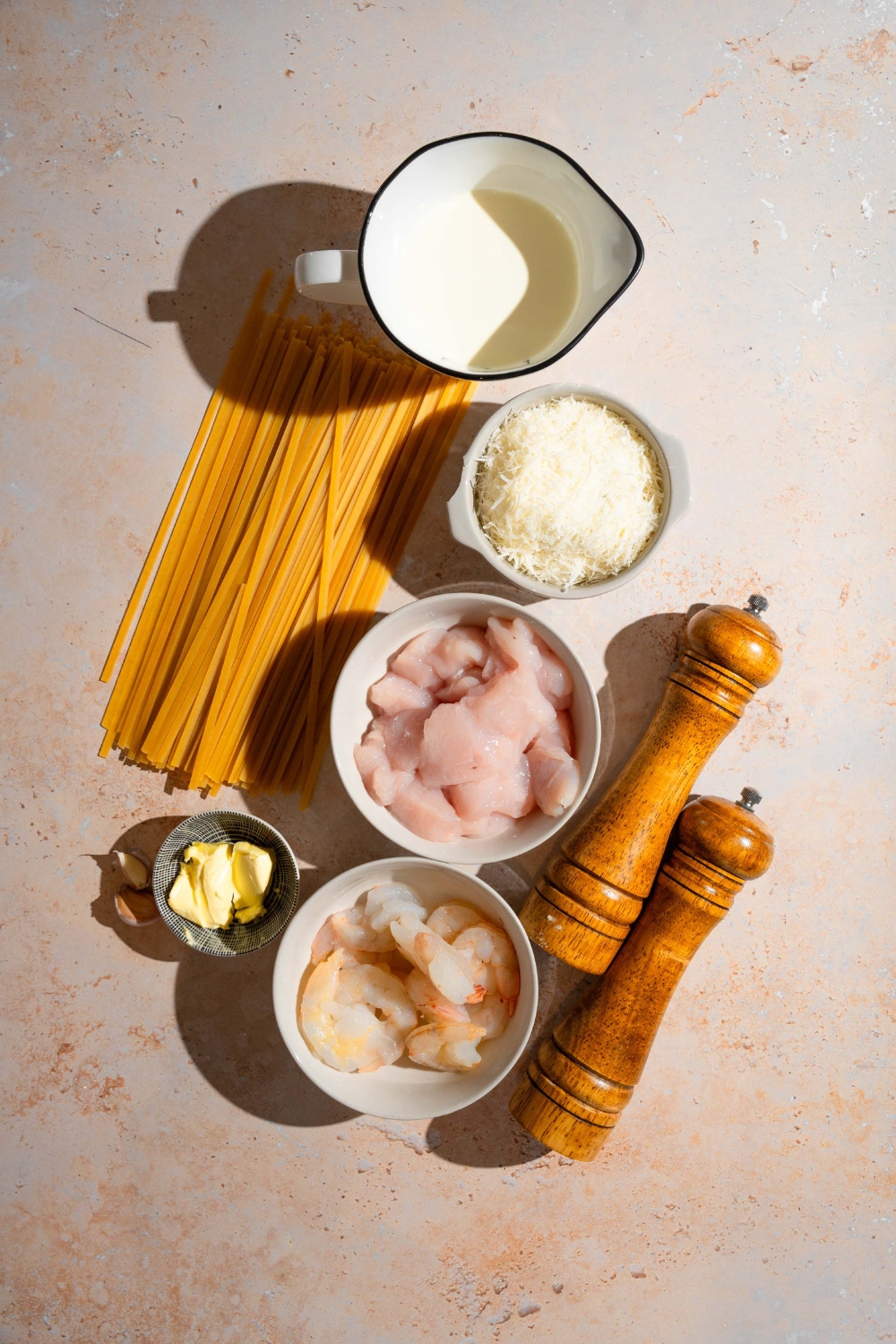 An overhead shot of several bowls in various sizes containing ingredients to make chicken and shrimp alfredo including chicken, shrimp, linguine pasta, cream, cheese, butter, salt, and pepper.