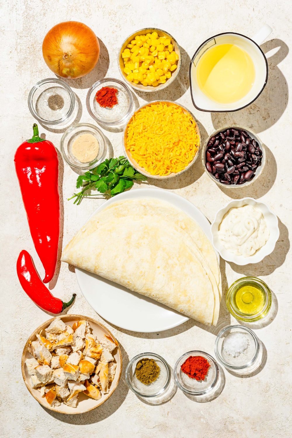 An overhead shot of several bowls in various sizes containing ingredients to make copycat Chili's southwestern egg rolls including egg roll wraps, shredded cheese, cubed chicken, black beans, red bell pepper, oil, corn, white onion, cilantro, and seasonings.