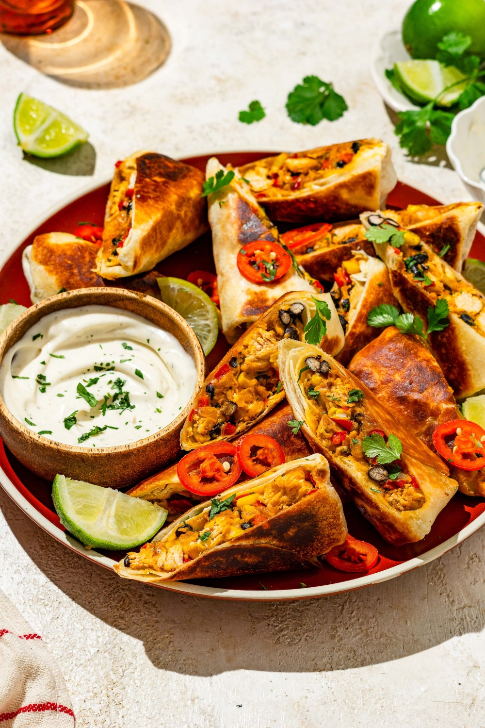 A plate of copycat Chili's southwestern egg rolls sliced in half and garnished with sliced peppers, fresh cilantro, and lime. The plate is served with a small bowl of sour cream. The plate is on a tan counter with lime wedges and cilantro.