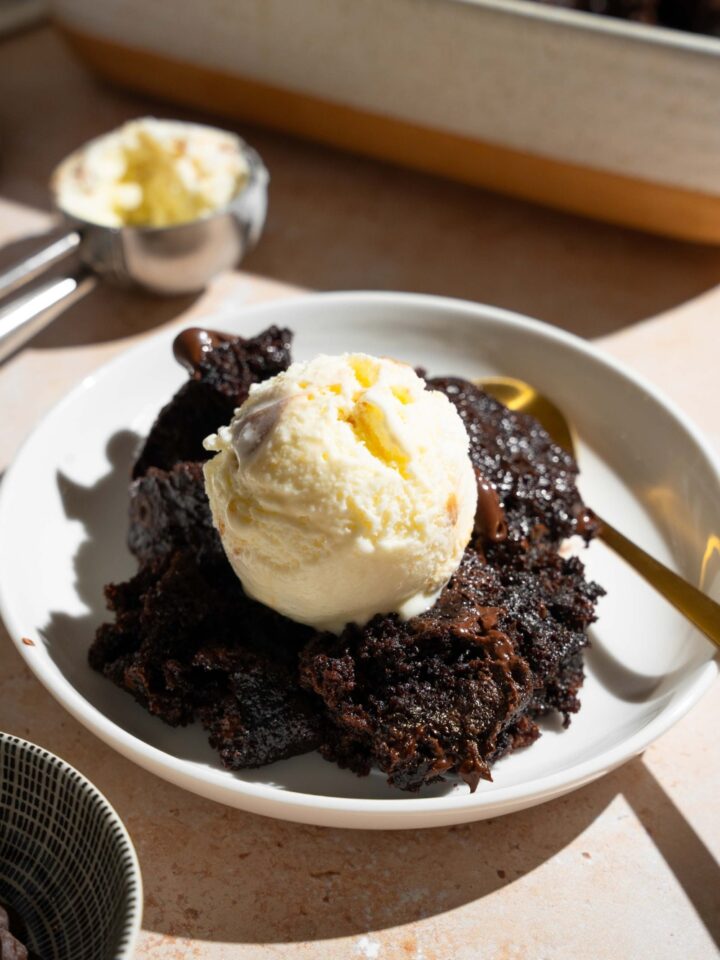 A white plate with a slice of chocolate dump cake topped with vanilla ice cream. There is a spoon on the plate. The plate is on a tan counter with a baking dish with dump cake.