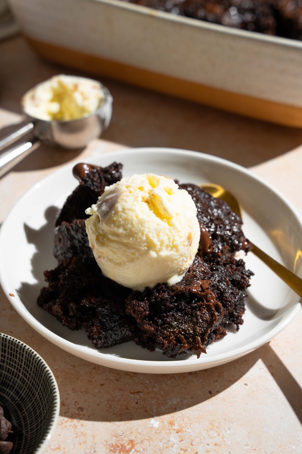 A white plate with a slice of chocolate dump cake topped with vanilla ice cream. There is a spoon on the plate. The plate is on a tan counter with a baking dish with dump cake.
