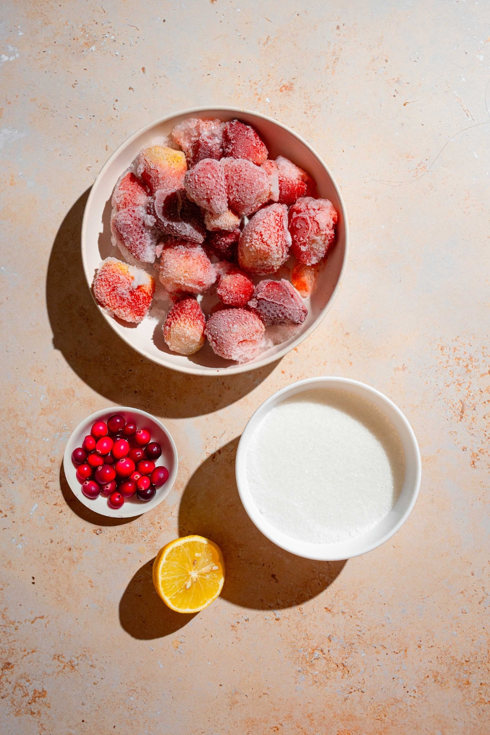 An overhead shot of several bowls in various sizes containing ingredients to make Christmas jam including frozen strawberries, sugar, cranberries, and lemon.