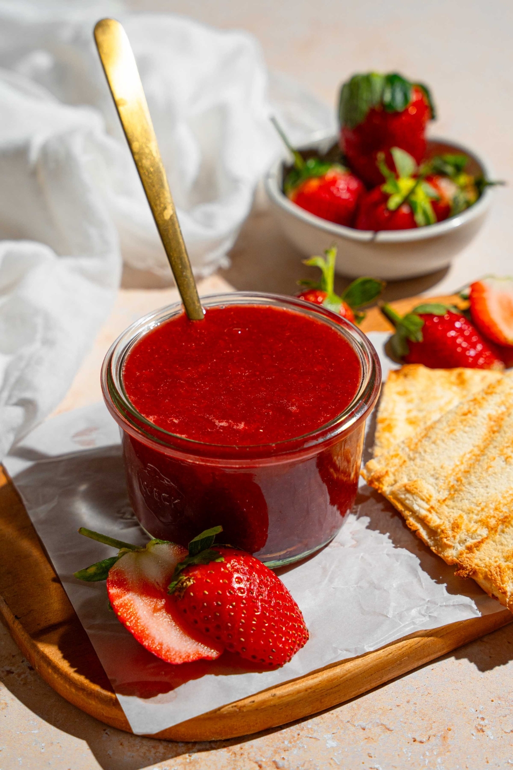 A jar of Christmas jam with a spoon in the jar. The jar is on a wooden board lined with parchment paper with a slice of toast and strawberries. The board is on a tan counter with a bowl of strawberries.
