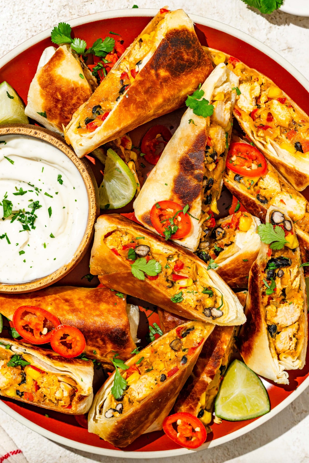 A plate of copycat Chili's southwestern egg rolls sliced in half and garnished with sliced peppers, fresh cilantro, and lime. The plate is served with a small bowl of sour cream.