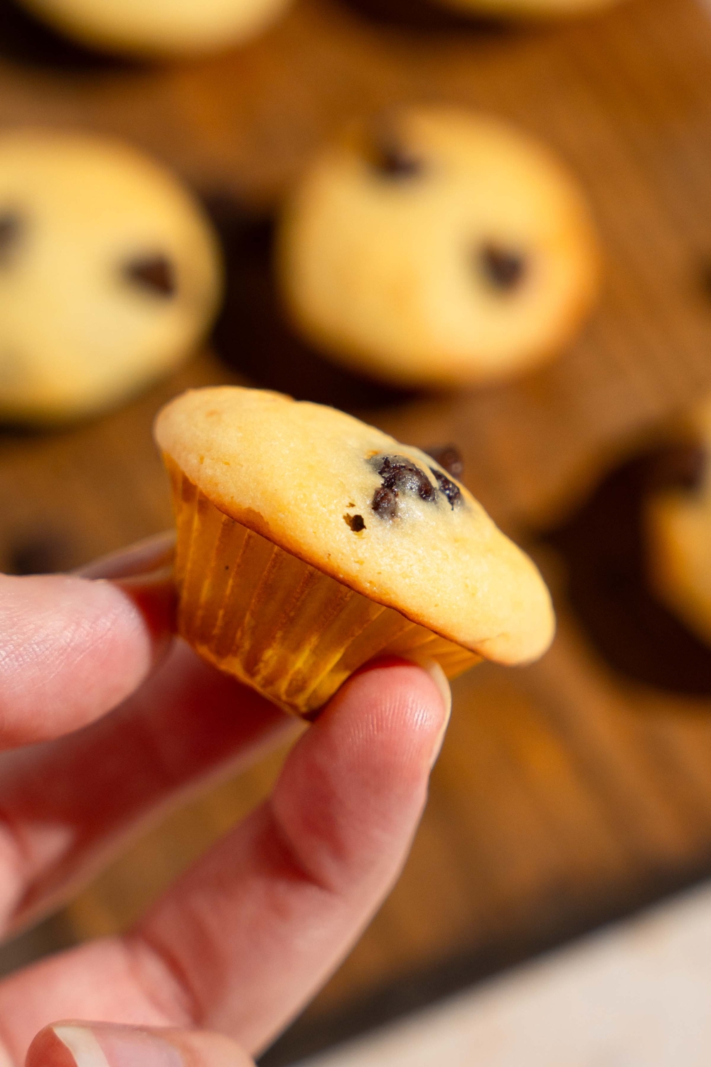 A hand holding a Little Bites chocolate chip muffin. There is a wooden board of muffins blurred in the background.