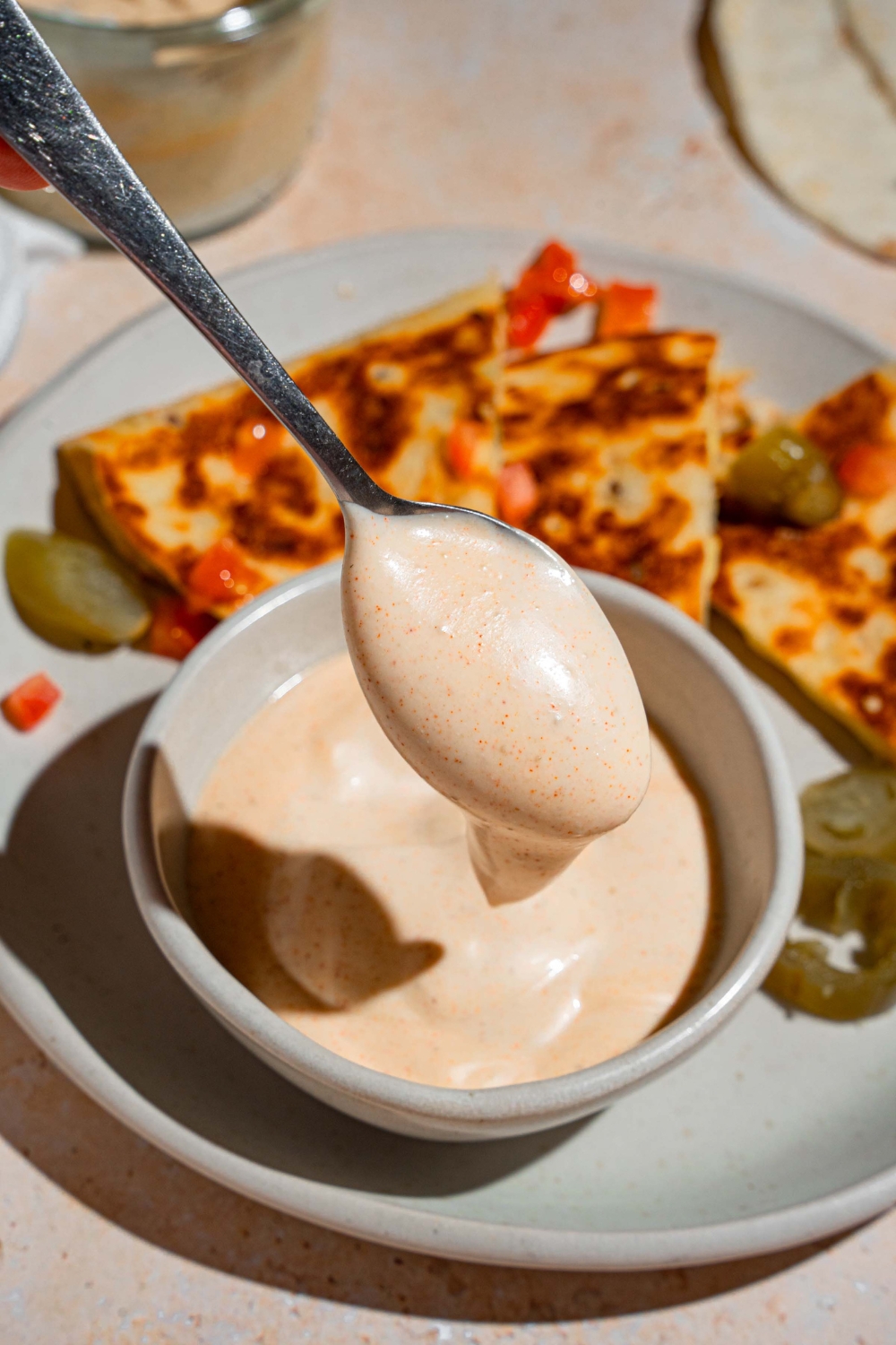 A spoon dipping into a bowl of Taco Bell quesadilla sauce. The bowl is on a white plate with a quesadilla garnished with peppers and pickled jalapenos. The plate is on a tan counter.
