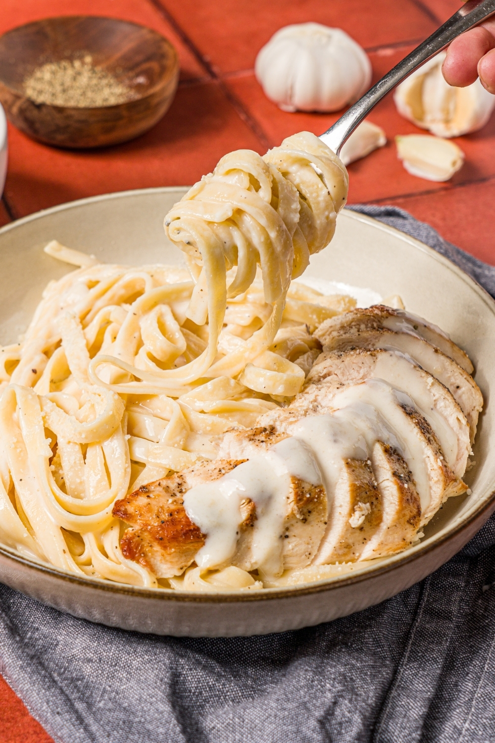 A bowl of chicken alfredo with a fork taking a bite of the fettuccine. The bowl is on a brick counter with a small bowl of pepper.