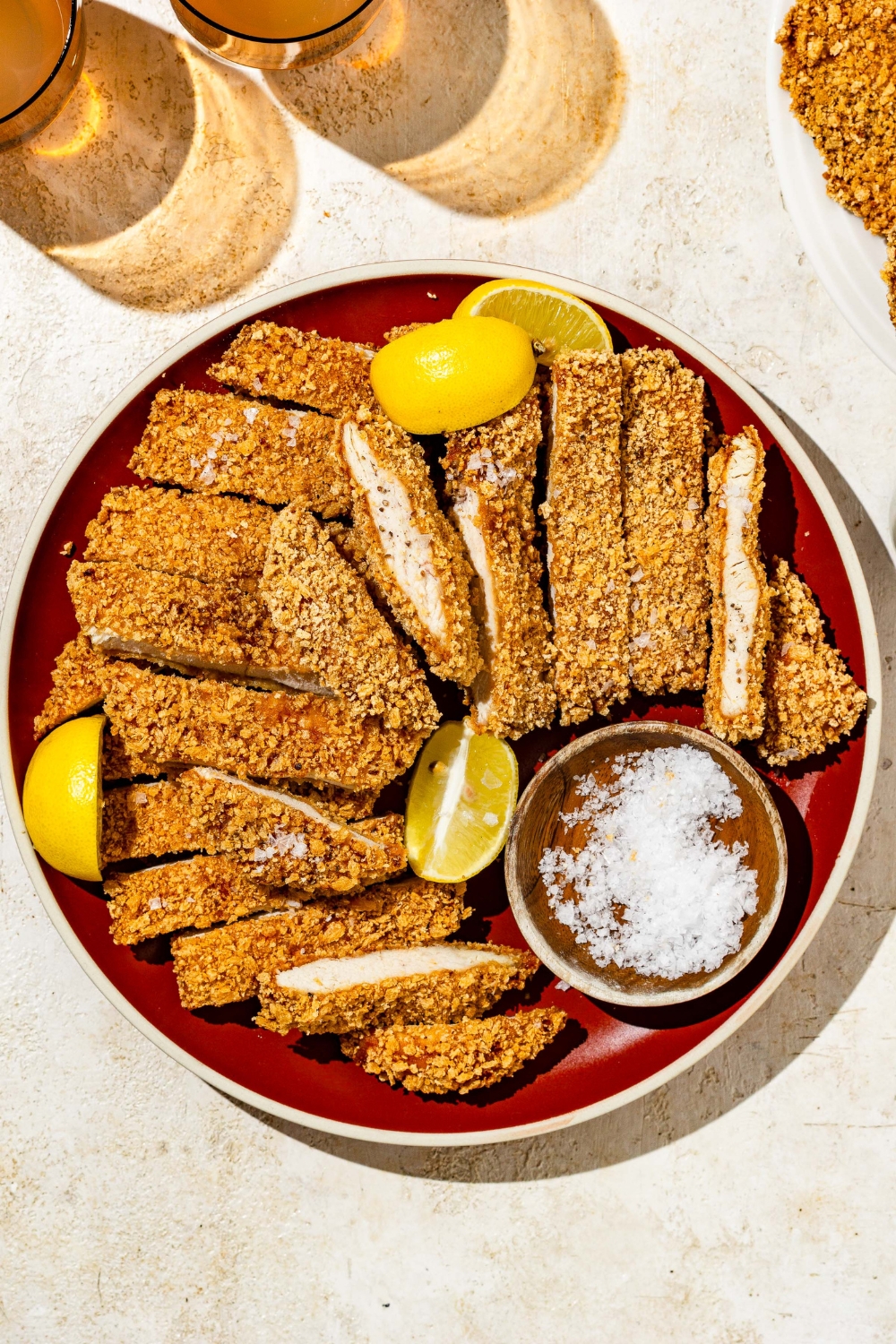 A plate of crispy chicken Katsu sliced in strips with a small ramekin of salt. The plate is garnished with lemon wedges. The plate is on a tan counter with glasses.