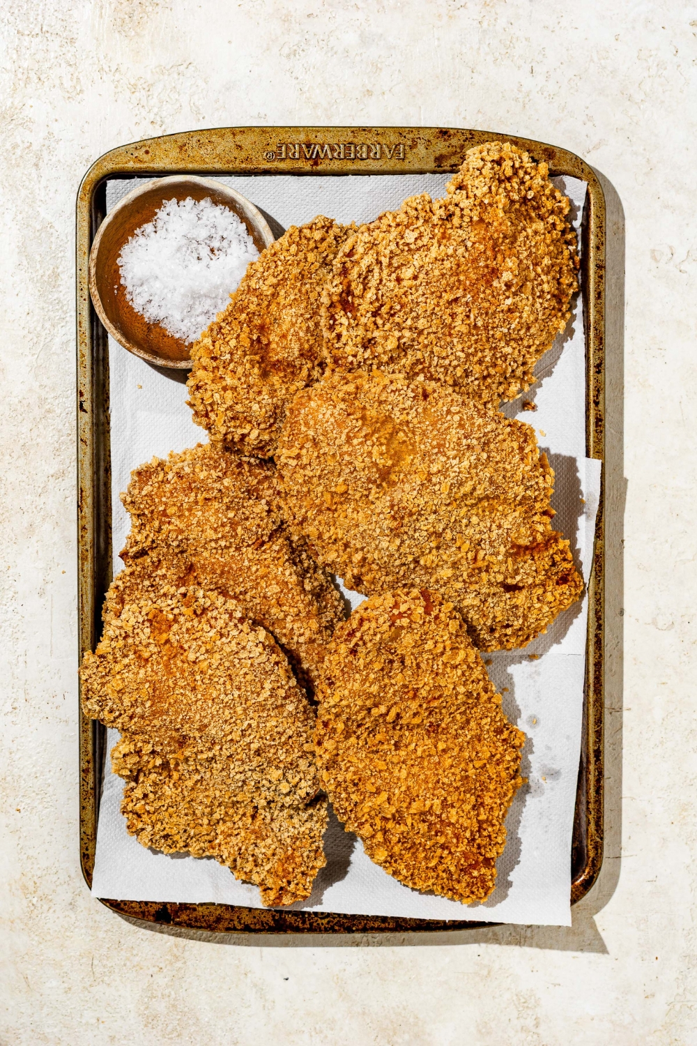 A baking sheet lined with parchment paper with several pieces of baked chicken Katsu. There is a small bowl of salt on the baking sheet.
