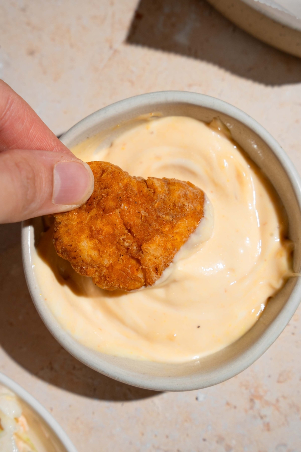 A hand dipping a homemade Chick Fil A nugget into a bowl of sauce. The bowl is on a tan counter.