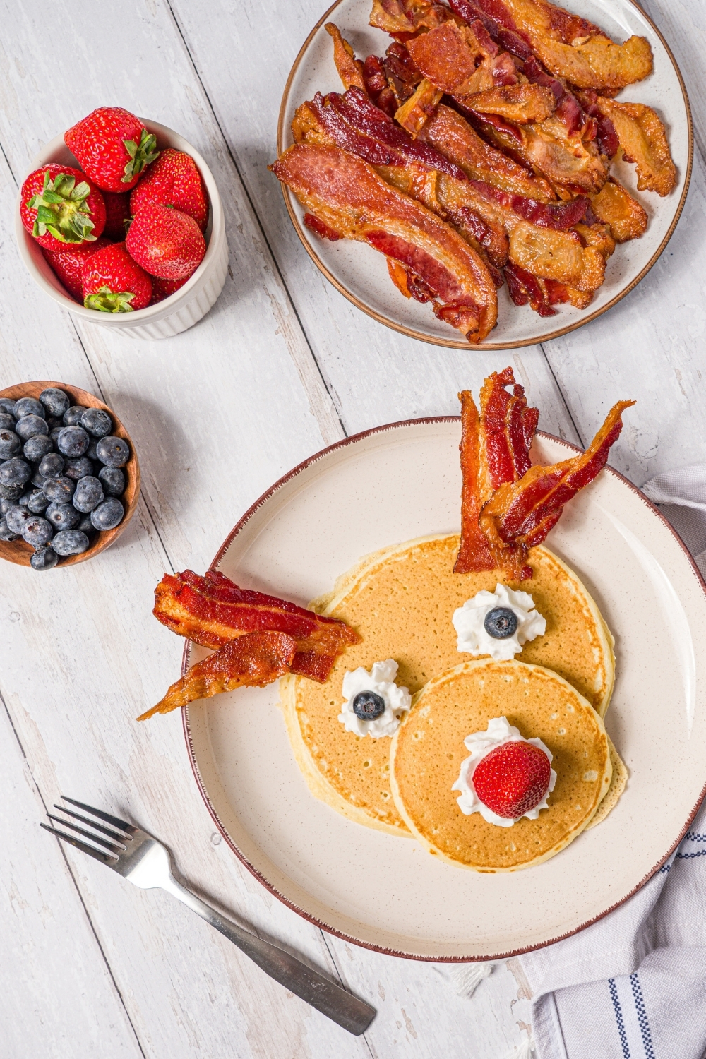 A white plate with reindeer pancakes topped with whipped cream, blueberry eyes, and a strawberry nose with bacon strips as antlers. The plate is on a wood counter with a plate of bacon and bowls of berries.