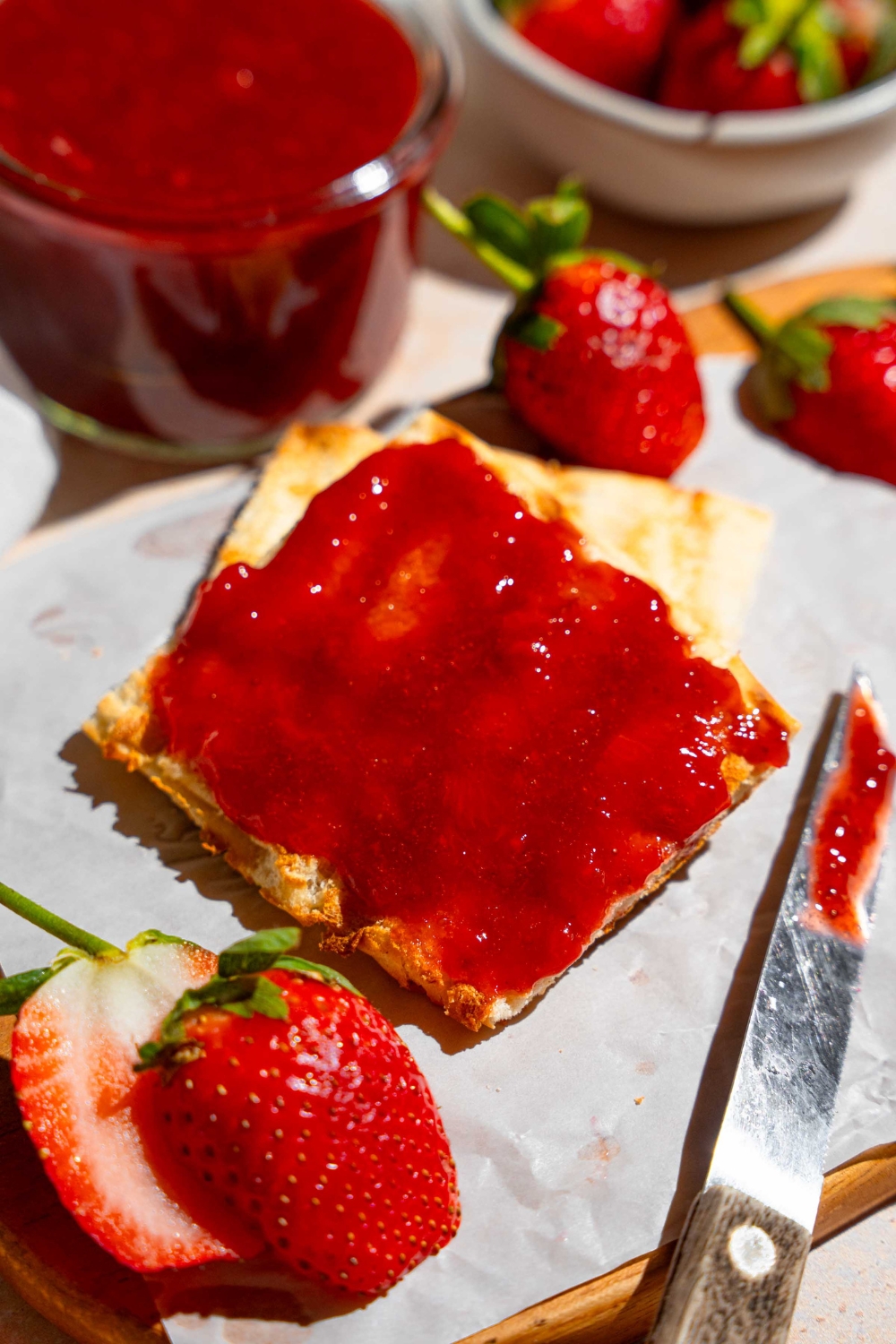 Two slices of toast with the top slice covered in Christmas jam. The toast is on a wooden board lined with parchment paper with a knife with jam and sliced strawberries. The board is on a tan counter with a jar of jam and bowl of strawberries.