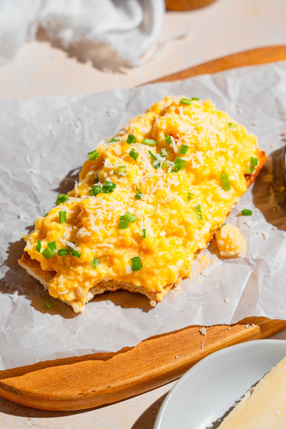 A thin slice toast topped with Gordon Ramsay scrambled eggs garnished with grated cheese and chives. The toast is on a wooden board lined with parchment paper and served with a ramekin of garnishes. There is a bite taken from the toast.