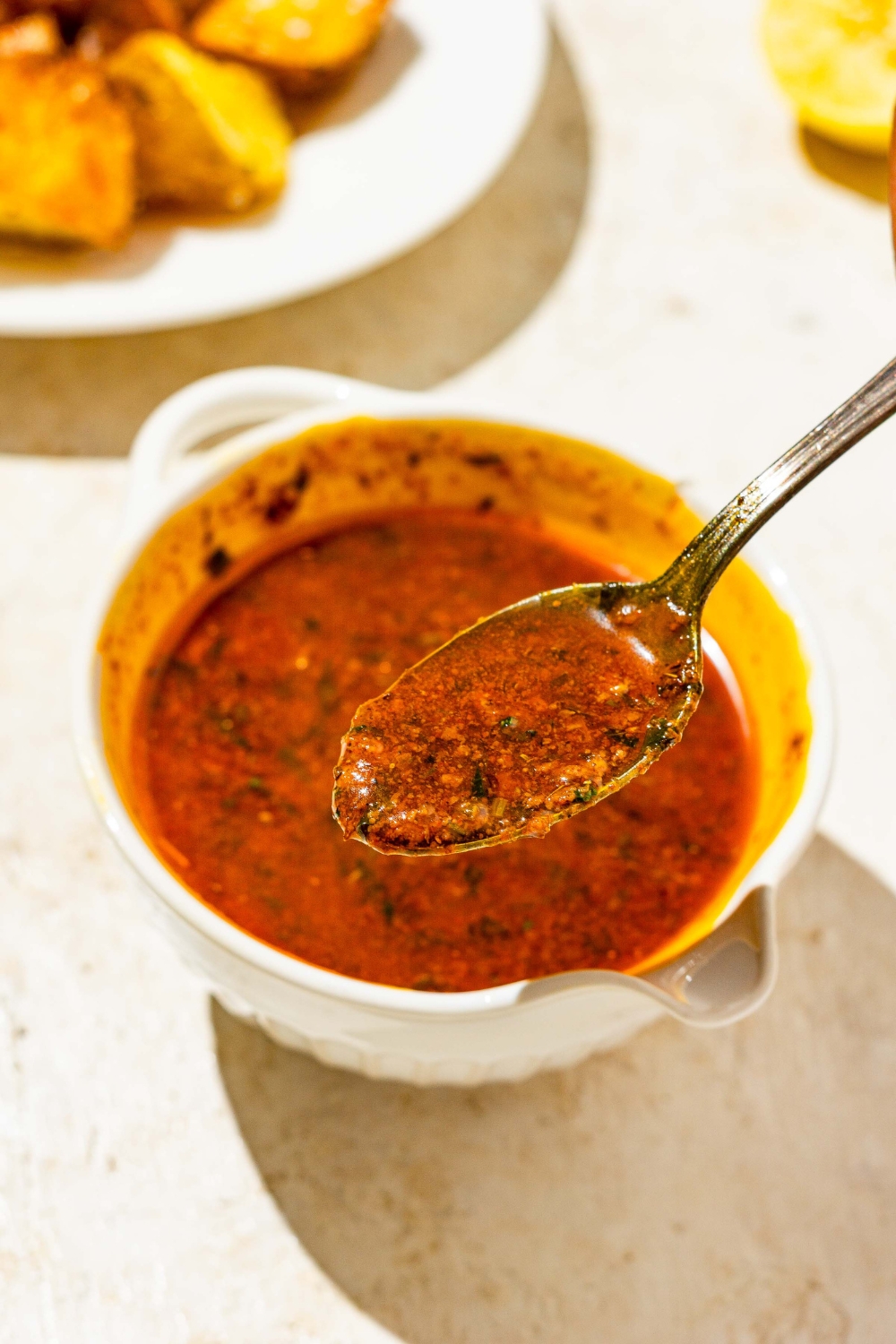 A close up of a spoon with cajun garlic butter sauce with a bowl of sauce blurred in the background. The bowl is on a tan counter.