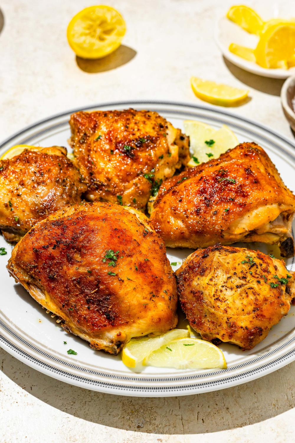 A plate of lemon pepper chicken thighs garnished with fresh parsley and lemon. The plate is on a tan counter with lemon.