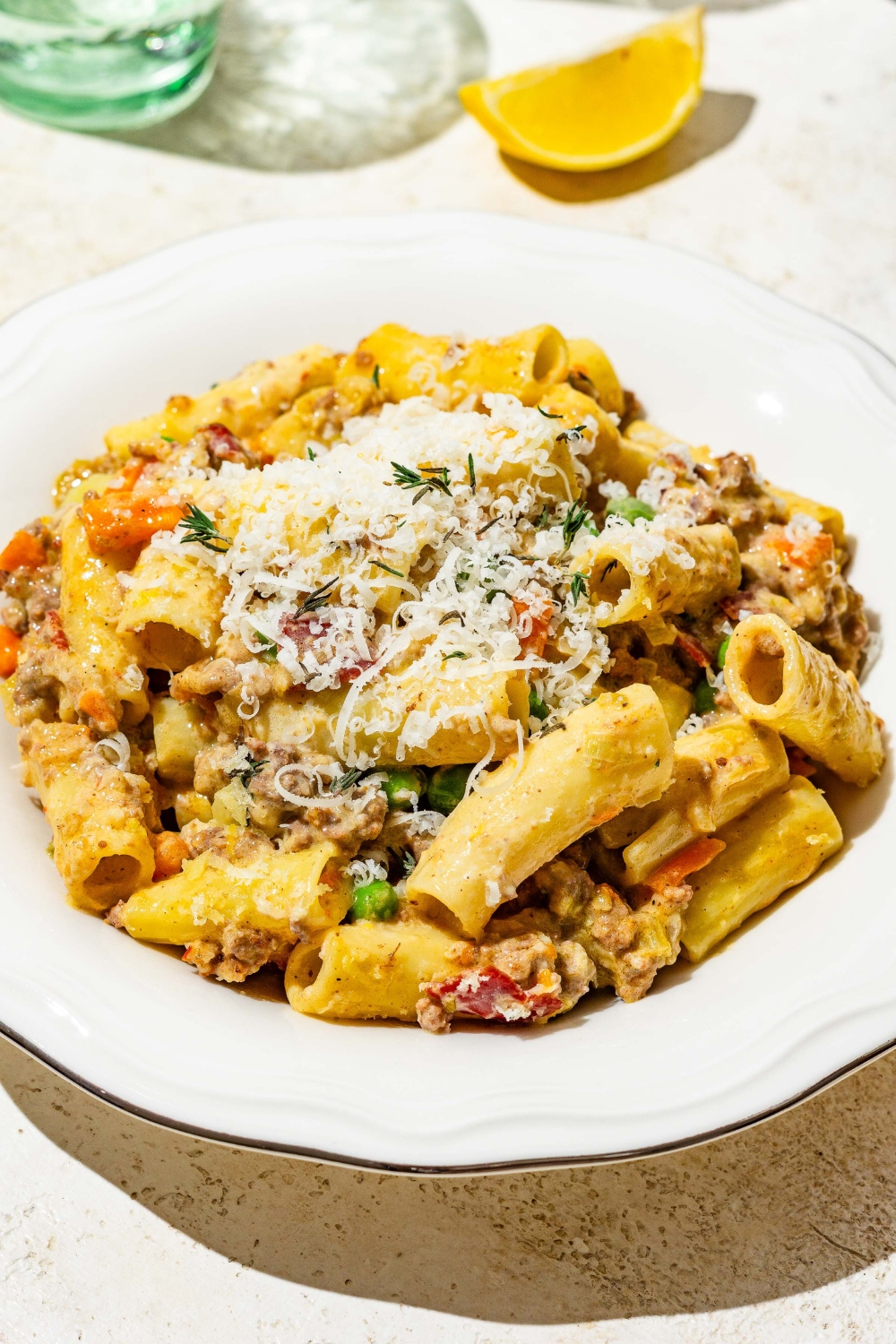 A white bowl with rigatoni pasta with white bolognese garnished with grated cheese. The bowl is on a tan counter with a lemon and glass of water.