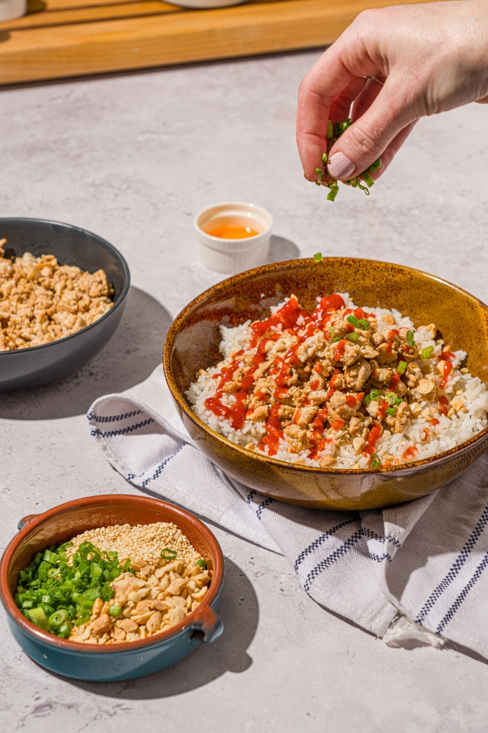 A bowl with ground turkey rice bowls with white rice, seasoned ground turkey, chopped peanuts, sliced green onions, sesame seeds, and drizzled with sriracha sauce. A hand is sprinkling green onion over the bowl. The bowl is on a white counter with a white striped napkin and a bowl of garnishes.