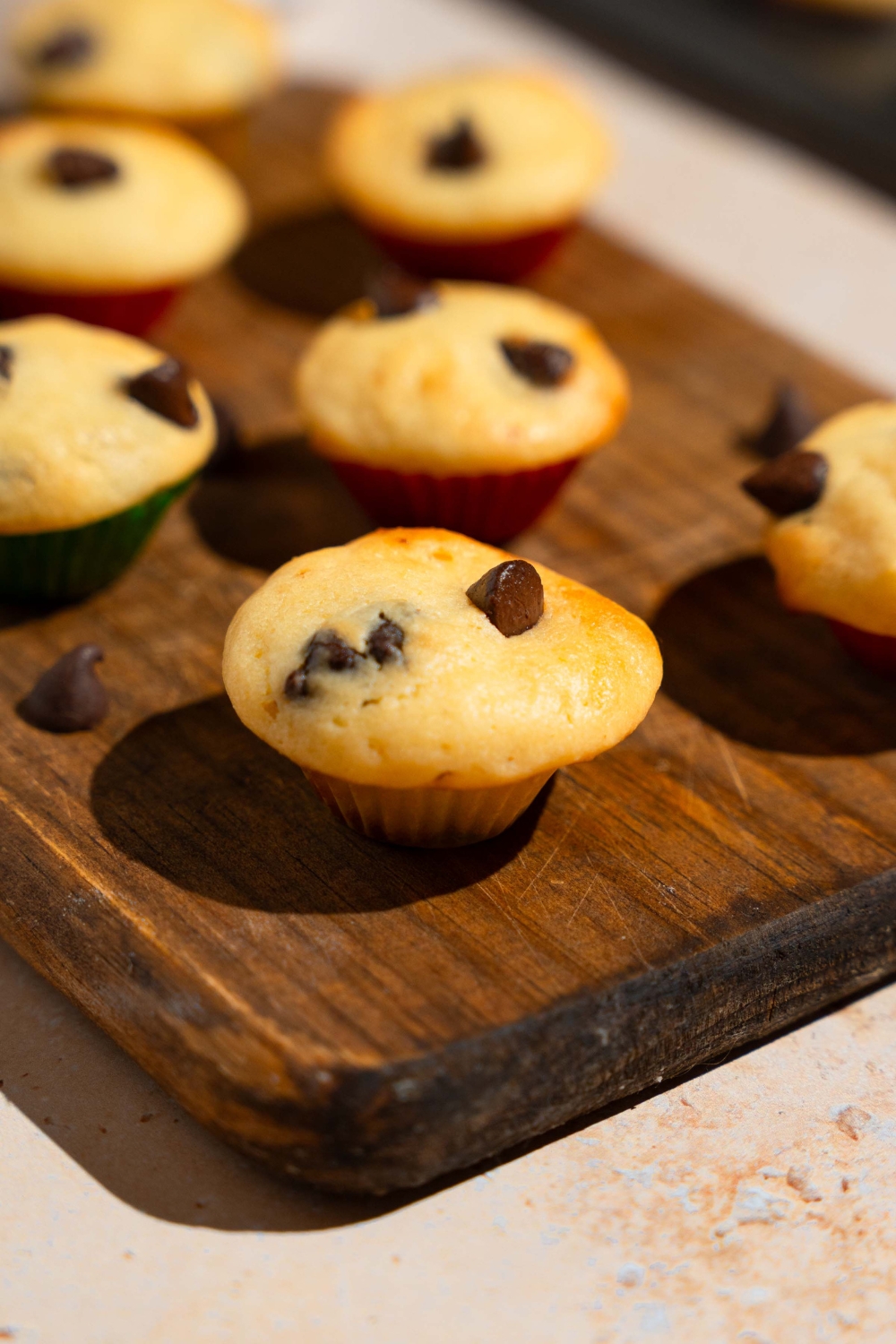 A wooden board with copycat Little Bites chocolate chip muffins. The board is garnished with chocolate chips. The board is on a tan counter.
