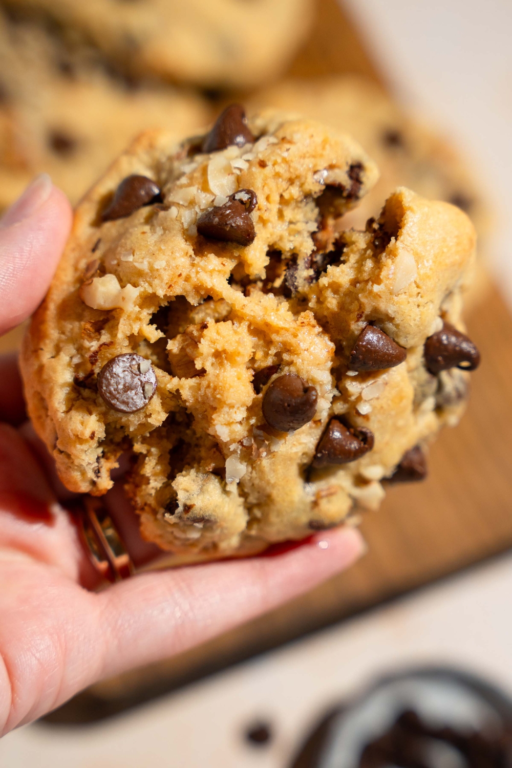 A close up of a hand holding Neiman Marcus chocolate chip cookie. There is a board of cookies blurred in the background.